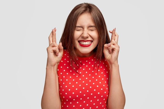 Pleased attractive European woman makes wish to win, raises hands with crossed fingers, waits for lottery results, closes eyes