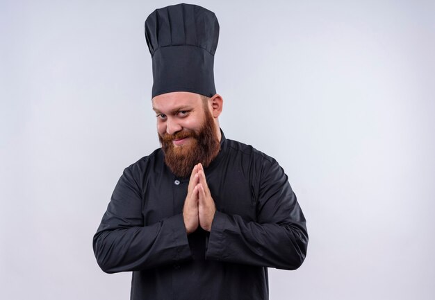 A pleased bearded chef man in black uniform holding hands together while looking on a white wall