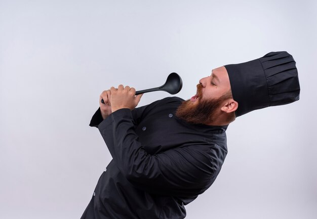 A pleased bearded chef man in black uniform singing to ladle using it like microphone on a white wall