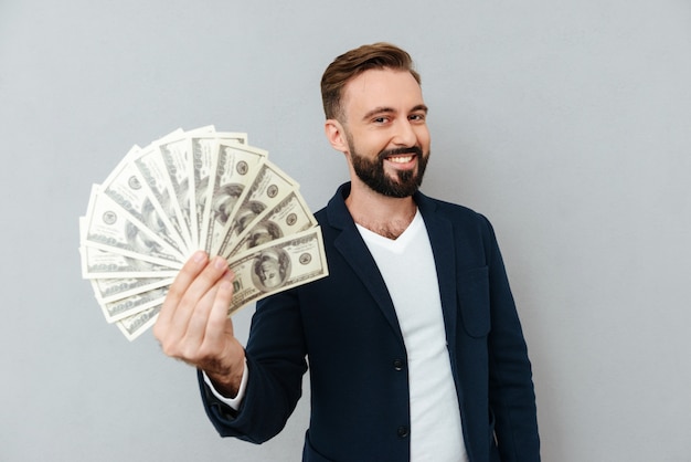 Pleased bearded man in busines clothes showing money and looking at the camera over gray 