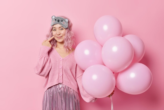 Pleased beautiful Asian woman with pink dyed hair points finger on face smiles pleasantly wears sleepmask jumper and skirt poses with balloons isolated over pink background. Festivity concept