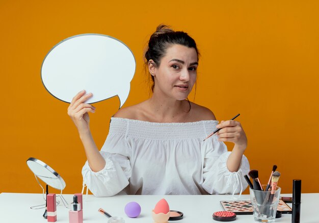 Pleased beautiful girl sits at table with makeup tools holds chat bubble and makeup brush isolated on orange wall