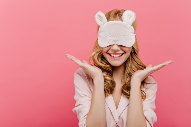 Free photo pleased beautiful young woman posing in eyemask. cheerful european girl in pajamas standing on pink wall in sleep mask in morning.