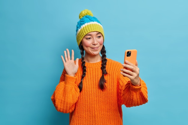 Pleased brunette pretty millennial girl waves palm greets friend online holds modern cellular makes video call wears warm knitted sweater and hat isolated over blue wall