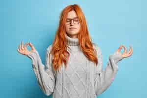 Free photo pleased calm redhead woman holds hand in yoga gesture for mental balance stands with closed eyes meditates to relax wears eyeglasses and sweater.