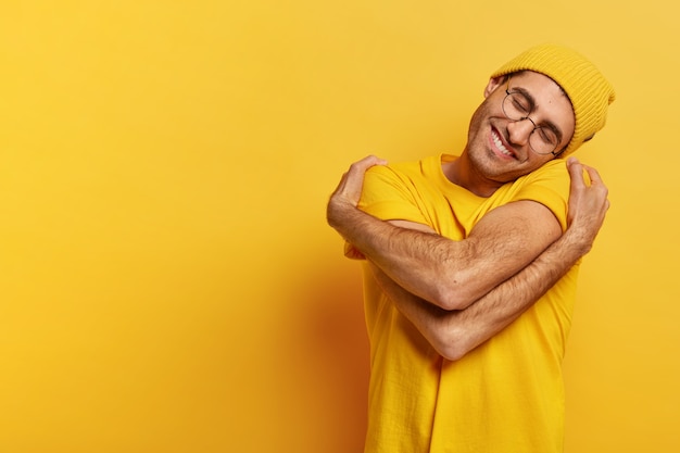 Free photo pleased caucasian man hugs himself, has high self esteem, tilts head, has toothy smile, wears casual yellow hat and t shirt