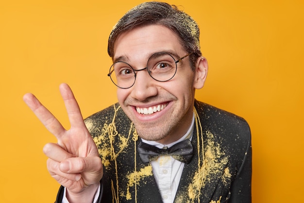 Free photo pleased cheerful adult man smiles gladfully being in good mood wears round spectacles and festive black formal suit enjoys celebration isolated over yellow studio wall hey stay always positive