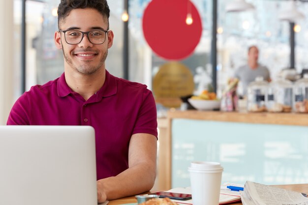 Pleased cheerful young man chats online with friends from abroad, sits in front of laptop computer, connected to 4G internet at coffee shop, wears optical glasses for good vision, likes his job