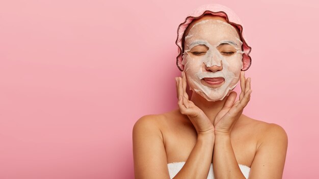 Pleased chinese woman enjoys cosmetic procedure, has natural paper face mask on cheeks, wrapped in towel, wears bathcap, has closed eyes, isolated on pink wall with free space for your advert