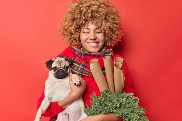 Free photo pleased curly haired woman bites lips keeps eyes closed giggles positively holds pedigree pug dog green spruce wreath and rolled paper for decor isolated over red background prepares for new year