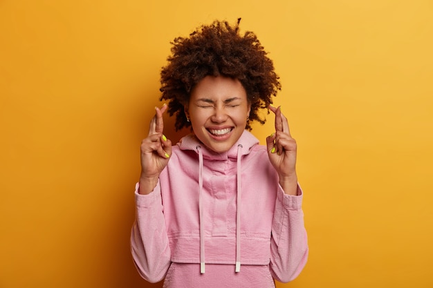 Free photo pleased curly woman crosses fingers, wishes fortune before exam, has great hope for better, smiles positively, closes eyes, wears velvet sweatshirt, poses over yellow wall, puts all efforts in praying