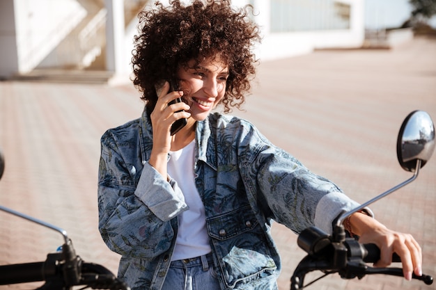 Pleased curly woman sitting on modern motorbike outdoors and talking by the smartphone