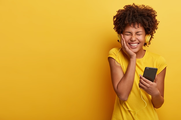 Pleased dark skinned woman feels great and happy, smiles broadly, holds smartphone connected to headphones, closes eyes from pleasure