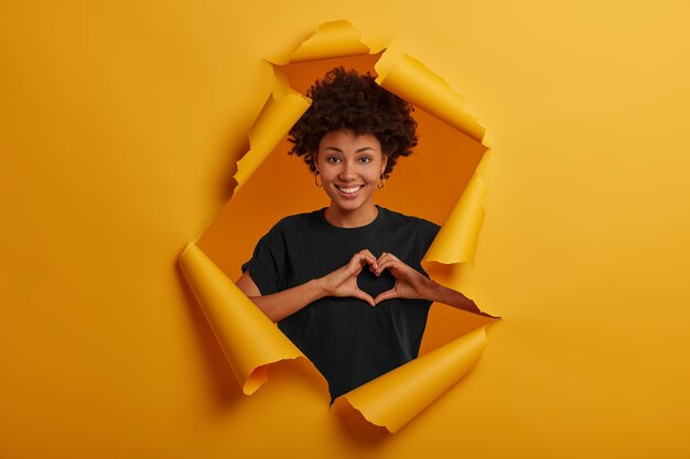 Pleased dark skinned young woman shows heart symbol, shapes love sign with hands, smiles happily, wears black t shirt and earrings