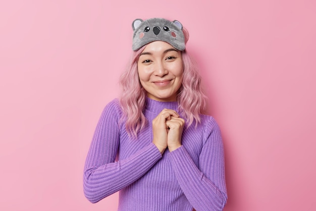 Free Photo pleased delighted asian woman with pink hair keeps hands together smiles gently glad to hear compliment wears sleepmask and blindfold isolated over pink background human facial expressions