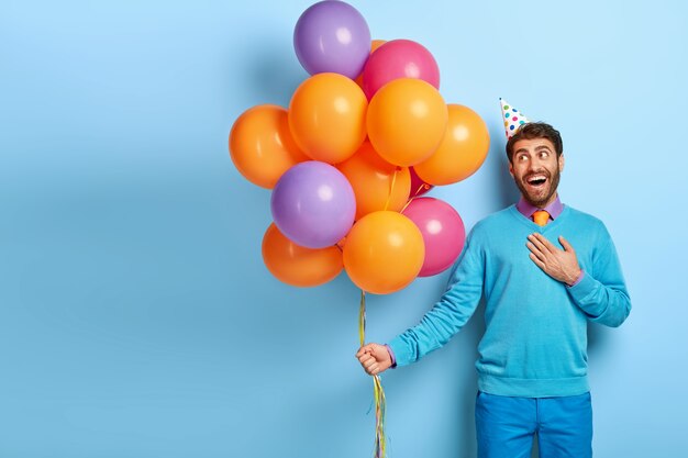 Pleased delighted guy with birthday hat and balloons posing in blue sweater