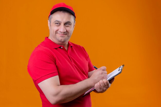 Pleased delivery man wearing red uniform and cap holding clipboard writing something looking aside with smile on face standing over orange background