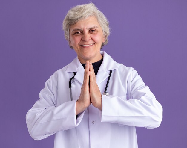 Pleased elderly woman in doctor uniform with stethoscope holding hands together 