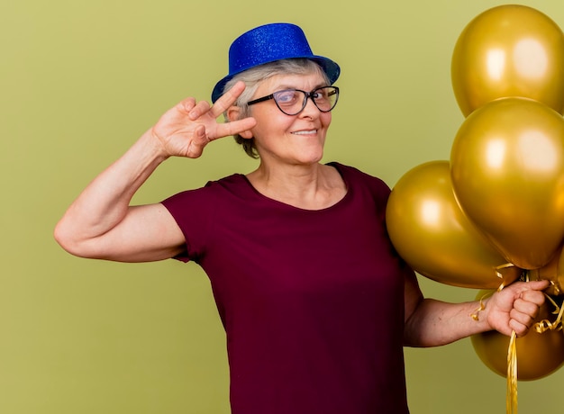 Pleased elderly woman in optical glasses wearing party hat stands with helium balloons gesturing victory