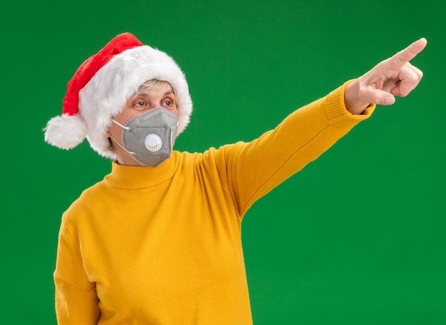 Free Photo pleased elderly woman with santa hat wearing medical mask looking and pointing at side isolated on green background with copy space