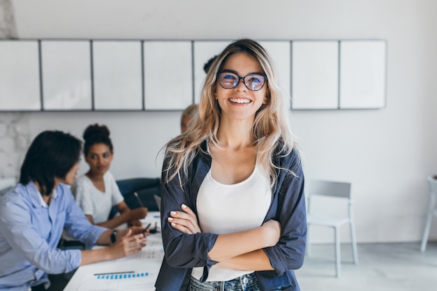 Free photo pleased female secretary in trendy glasses posing in office after meeting with colleagues. indoor portrait of stylish businesswoman with asian and african workers.