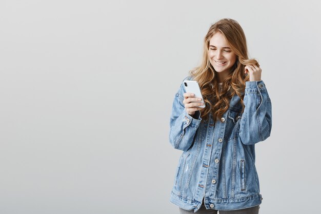 Pleased flirty blond girl smiling at smartphone intrigued