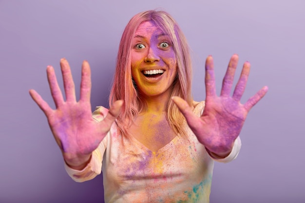 Free photo pleased funny young woman stretches both palms smeared with colored dry powder, happy facial expression, has fun with friends during holi festival, isolated against purple wall.