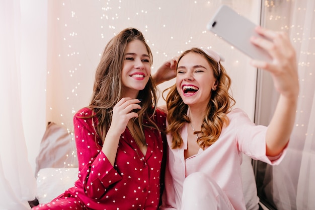 Pleased girl in pink pajamas having fun in her room with best friend. Cheerful young lady making selfie in morning with sister.