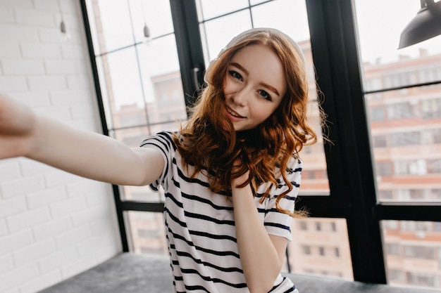 Free Photo pleased girl with ginger curly hair taking picture of herself. indoor shot of pretty woman making selfie.