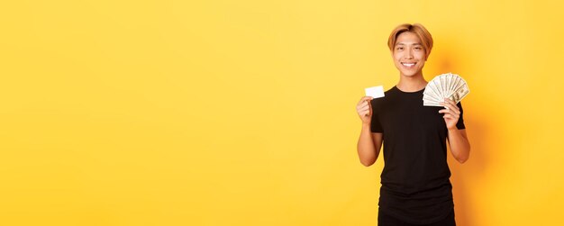 Pleased handsome asian guy showing money and credit card smiling happy standing yellow background