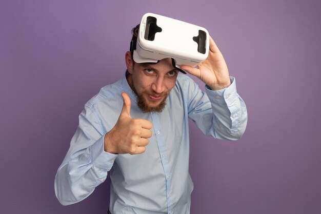 Pleased handsome blonde man holds vr headset and thumbs up isolated on purple wall