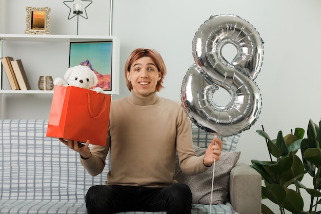 Free Photo pleased handsome guy on happy women day holding number eight balloon with gift bag sitting on sofa in living room