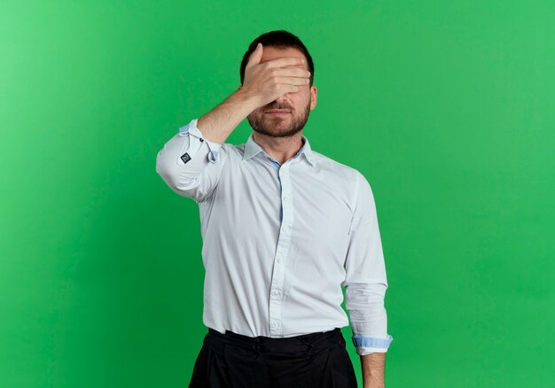 Pleased handsome man closes eyes with hand isolated on green wall