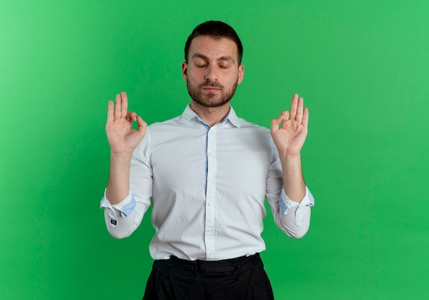 Pleased handsome man pretends to meditate with closed eyes isolated on green wall