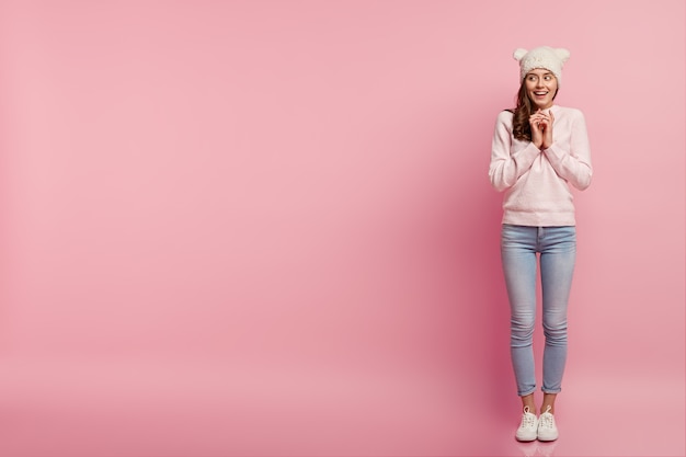 Pleased happy woman keeps hands together, looks aside with intention to do something, wears white hat with ears