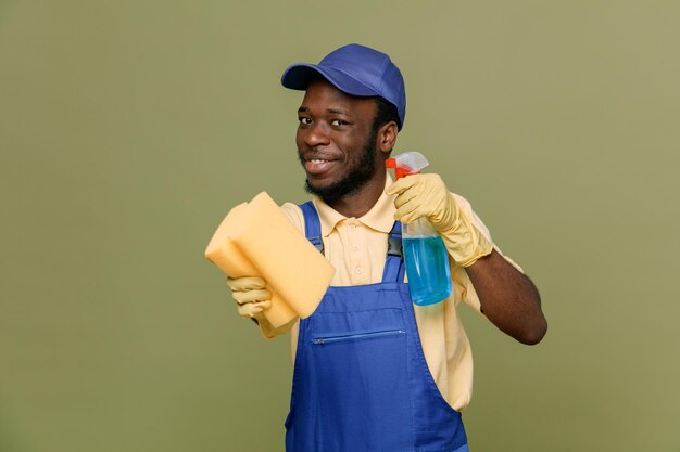 Pleased holding cleaning agent with sponge young africanamerican cleaner male in uniform with gloves isolated on green background