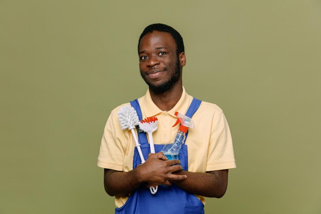 Pleased holding cleaning tools young africanamerican cleaner male in uniform with gloves isolated on green background