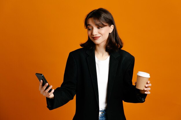 Pleased holding coffee cup and phone young beautiful female wearing black jacket isolated on orange background