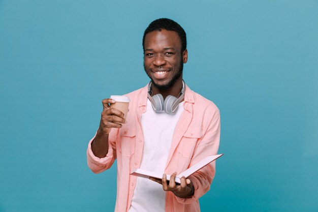 Pleased holding coffee cup with book young africanamerican guy wearing headphones on neck isolated on blue background