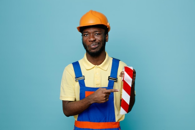 Pleased holding and points duct tape young african american builder in uniform isolated on blue background