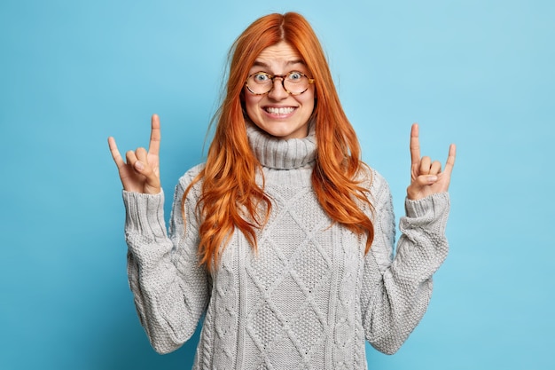 Pleased joyous redhead woman in spectacles shows rock n roll hand sign devil horn gesture smiles broadly shows white teeth wears winter sweater.