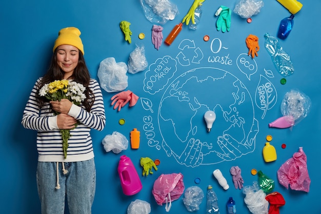 Free Photo pleased korean woman satisfied to get bouquet, holds white and yellow flowers, stands against drawn planet and plastic trash around on blue wall, cleans nature from pollution.