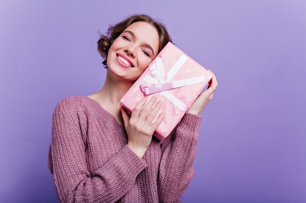 Pleased lady with glamorous makeup posing with new year gift on purple wall. Dreamy short-haired girl standing with eyes closed , holding birthday present.