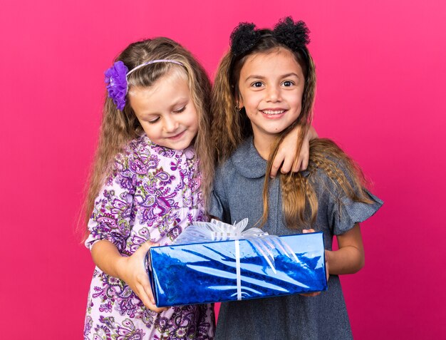 pleased little pretty girls holding gift box together isolated on pink wall with copy space