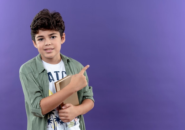 Pleased little schoolboy holding book and points at side isolated on purple wall with copy space