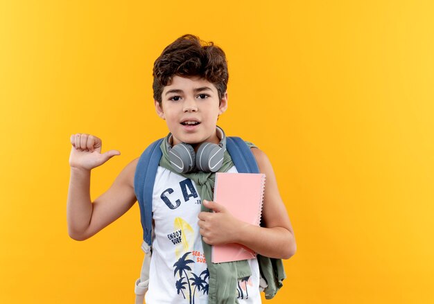 Pleased little schoolboy wearing back bag and headphones holding notebook isolated on yellow background