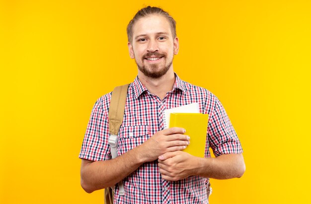 Pleased looking camera young guy student wearing backpack holding book isolated on orange wall