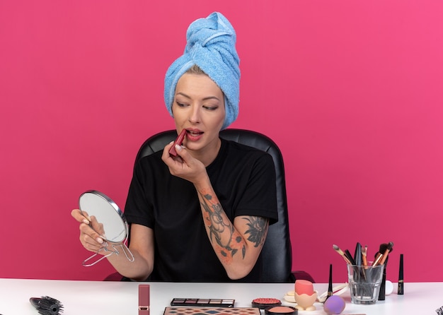 Pleased looking at mirror young beautiful girl sits at table with makeup tools wrapped hair in towel applying lipstick isolated on pink wall