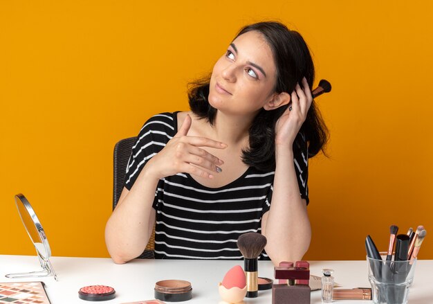 Pleased looking side young beautiful girl sits at table with makeup tools holding powder brush isolated on orange wall