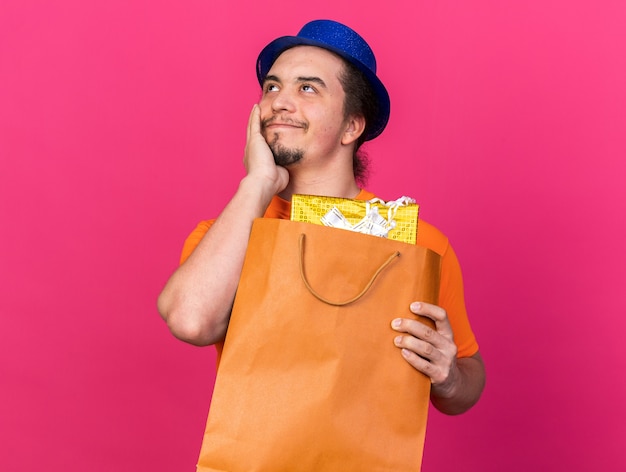 Pleased looking side young man wearing party hat holding gift bag putting hand on cheek isolated on pink wall